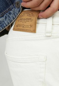 Białe dżinsy z denimu z skórzaną metką marki, na której znajduje się logo "Jack & Jones", umieszczoną na tylnej kieszeni, podkreślając ręcznie robione towary.