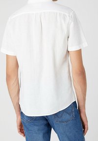 Biała koszula z krótkim rękawem, uszyta z lekkiego materiału, z standardowym kołnierzykiem i detalem yoke na plecach. Połączona z ciemnoniebieskimi dżinsami.