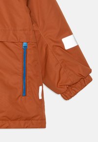 Brun jakke med blå lynlås og elastiske manchetter, med en sidelomme og reflekterende detaljer. Glat stofstruktur med et enkelt design.