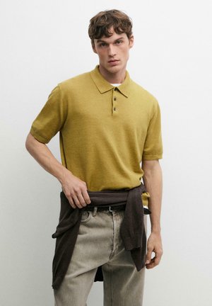 Młody mężczyzna ubrany w żółte polo z krótkim rękawem, jasnoszare jeansy i ciemny sweter związany wokół talii, stoi przy białej ścianie.