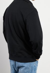 Persona con una camiseta de manga larga negra lisa y jeans azules, con las manos en los bolsillos, de espaldas contra un fondo blanco.