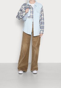 Henkilö, joka pukeutunut leveälahkeisiin ruskeisiin housuihin, valkoisiin tennareihin ja siniseen sekä ruudulliseen tilkkutäkki-paidan, pitäen vaaleansinistä olkalaukkua.