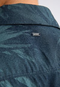 Camisa de color azul marino con un patrón texturizado; presenta un cuello y una pequeña etiqueta de logotipo plateado cerca del escote, resaltando su tejido ligero.