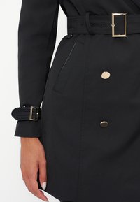 Gros plan sur un trench noir avec des boutons dorés, une ceinture assortie avec une boucle dorée et une main avec un vernis à ongles clair.