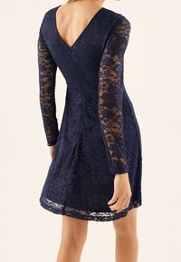 Femme portant une robe en dentelle florale bleu marine avec de longues manches et un dos en V profond, se tenant devant un fond clair uni.