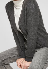 Donkergrijze gebreide cardigan met een v-hals en ribbeltextuur, voorzien van knoopsluiting en lange mouwen, gedragen over een witte coltrui.