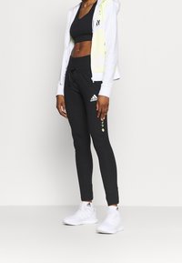 Leggings athlétiques noirs avec logo Adidas, assortis à un sweat à capuche court blanc avec des accents néon et un soutien-gorge de sport noir. Baskets blanches.