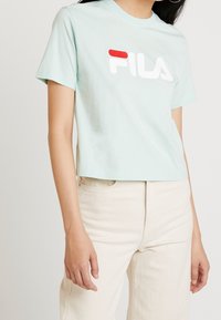 Camiseta corta de color azul claro hecha de algodón, con un gran logo de FILA en blanco y rojo en la parte delantera. Mangas cortas y un diseño de corte relajado.