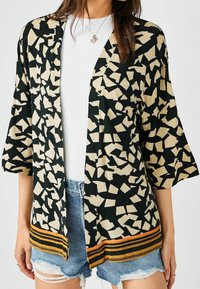 Schwarzer und beiger Kimono mit einem abstrakten Muster, der breite Ärmel und einen gestreiften Saum in Orange und Gelb aufweist, kombiniert mit Jeansshorts.