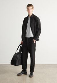 Veste noire sur un polo gris, pantalon noir et baskets noires. Le modèle tient un sac de sport noir, se tenant contre un fond clair.