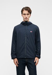 Νεαρός άνδρας με ναυτική ζακέτα με φερμουάρ και καπέλο που έχει ένα μικρό κόκκινο, λευκό και μπλε λογότυπο στο στήθος, συνδυασμένη με σκούρα τζιν, στέκεται μπροστά σε άσπρο φόντο.