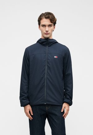 Jeune homme portant une veste à capuche zippée bleu marine avec un petit logo rouge, blanc et bleu sur la poitrine, associé à un jean sombre, se tenant devant un fond blanc.