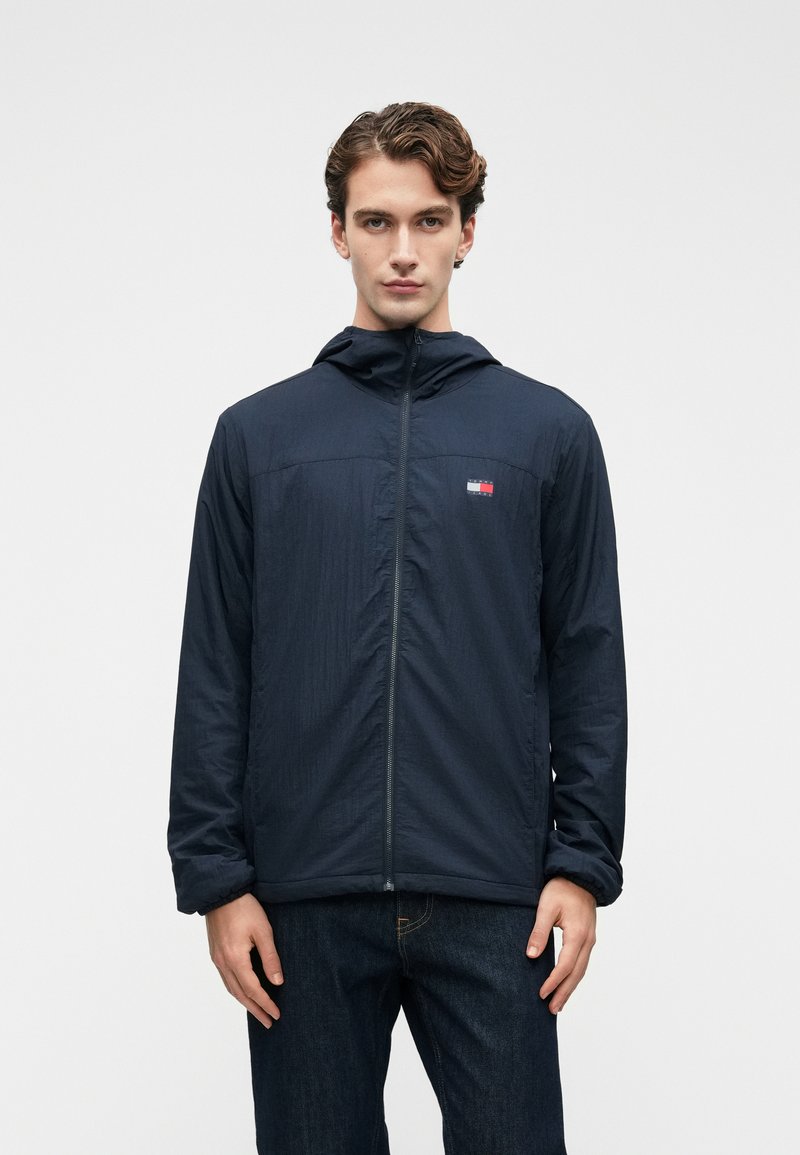 Νεαρός άνδρας με ναυτική ζακέτα με φερμουάρ και καπέλο που έχει ένα μικρό κόκκινο, λευκό και μπλε λογότυπο στο στήθος, συνδυασμένη με σκούρα τζιν, στέκεται μπροστά σε άσπρο φόντο.