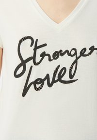Camiseta blanca de cuello en V con letras en negro que dicen "Amor más fuerte", adornada con pequeñas cuentas negras para textura y detalle.