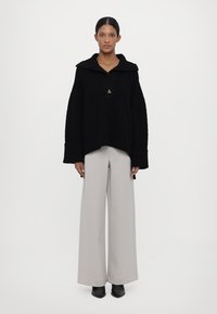 Black knitted oversized sweater with a wide collar, featuring a small gold triangle accent. Paired with light grey wide-leg trousers and black shoes.
