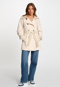 Manteau beige à double boutonnage, taille cintrée et poches latérales. Présente des boutons en métal argenté et un détail à ceinture.