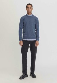 Blauer Strickpullover mit Rundhalsausschnitt, kombiniert mit einem hellblauen Hemd mit Kragen, schwarzen Hosen und schwarzen Schuhen. Glatte Textur, taillierte Passform.