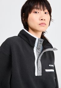 Schwarzer Fleece-Pullover mit hohem Kragen, grauen geknöpften Akzenten und weißen Details. Verfügt über eine strukturierte, weiche Oberfläche und eine Brusttasche.