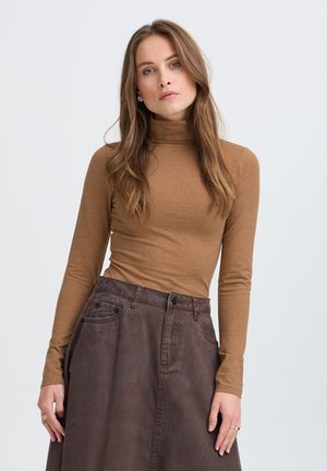 Femme aux longs cheveux bruns portant un col roulé marron et une jupe taille haute marron foncé, debout devant un fond clair uni.