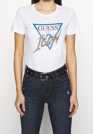 T-shirt blanc avec un logo triangulaire bleu portant les inscriptions "GUESS" et "ICON" en strass. Associé à un jean en denim bleu foncé et une ceinture noire.