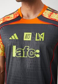 Mørkegrå atletisk skjorte med lyse orange og mønstrede ærmer. Har gule logoer: "lafc," "NTS," og Adidas. Tekstureret stof med mesh-detaljer.