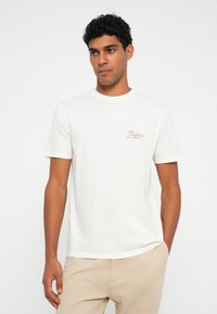 Camiseta de algodón blanca de manga corta; presenta un pequeño logo marrón en el lado izquierdo del pecho. Textura suave y cuello redondo clásico.