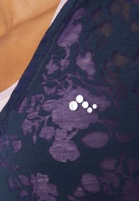 Tmavě modrý top s fialovým květinovým vzorem, s výstřihem do V a jemným stříbrným logem na levé straně. Měkká textura.