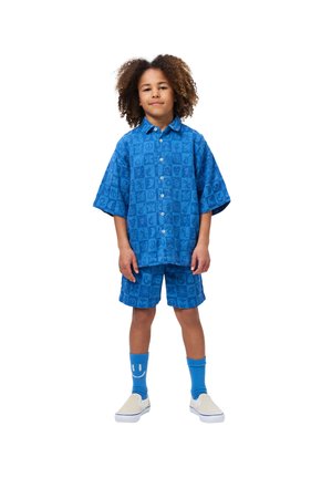 Modrá košile s krátkým rukávem a odpovídající šortky s texturovaným vzorem mírových znaků a srdcí. Stylizováno s modrými ponožkami a béžovými klouzavými botami.