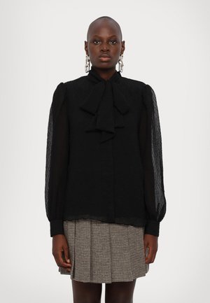 Blusa nera con tessuto testurizzato, caratterizzata da un fiocco al collo, maniche puff trasparenti e chiusura con bottoni, abbinata a una gonna a motivo houndstooth.