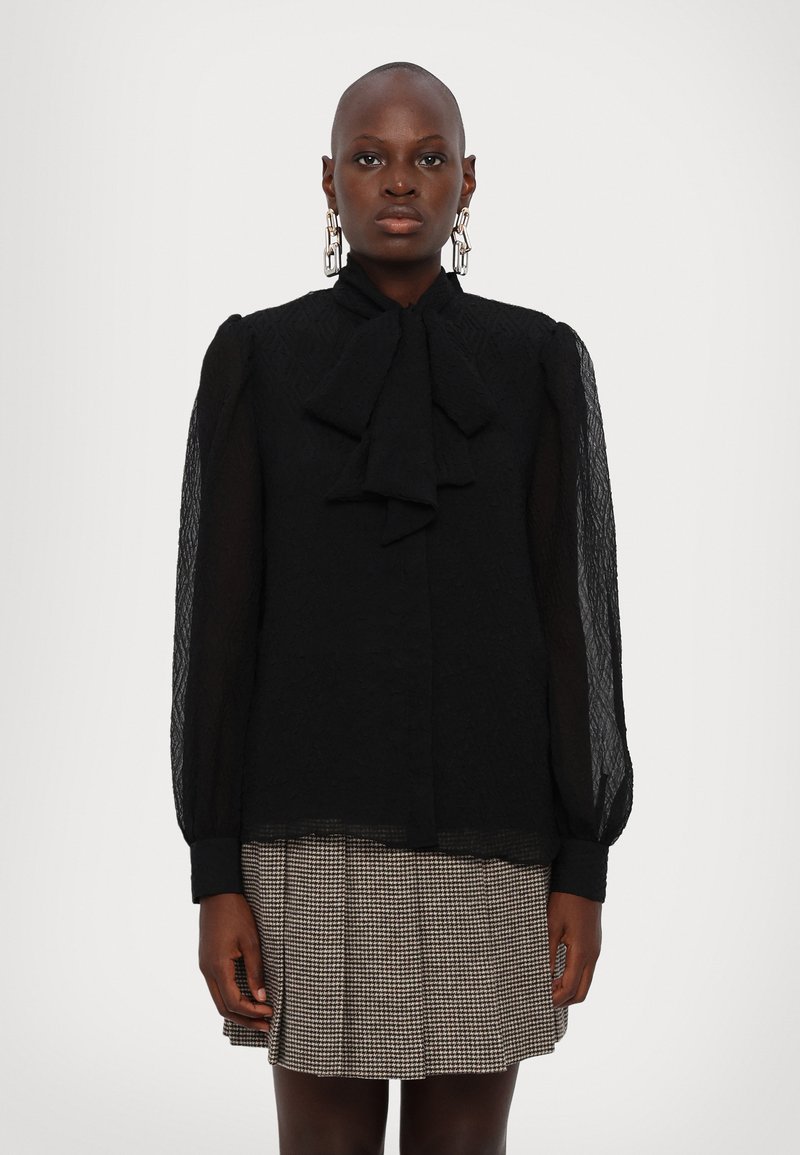 Schwarze Bluse aus strukturiertem Material mit einer Schleife am Kragen, durchsichtigen Puffärmeln und Knopfverschluss, kombiniert mit einem Hahnentrittmuster-Rock.