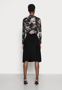 Blouse noire à fleurs à manches longues associée à une jupe plissée noire de longueur genoux. Le mannequin porte des mules à talons noires. Fond blanc uni.