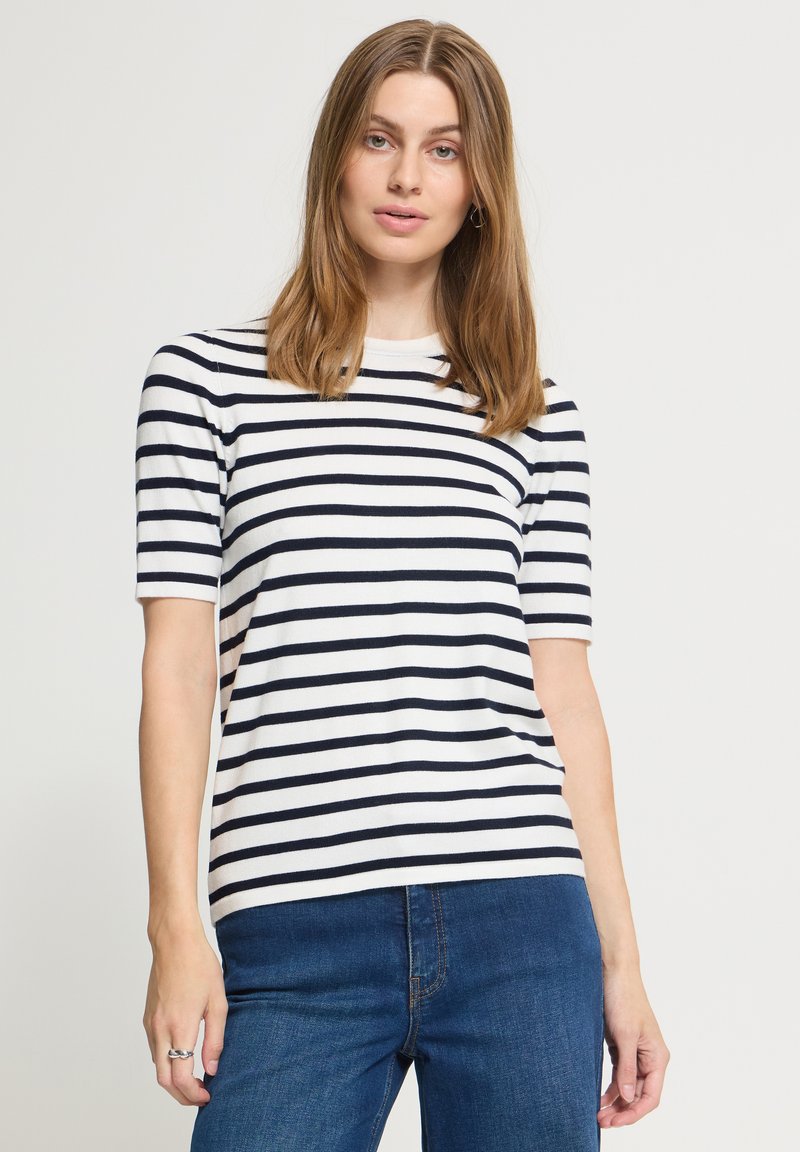 Femme aux cheveux lisses châtain clair portant une chemise blanche à manches courtes avec des rayures horizontales noires et un jean bleu sur un fond uni.