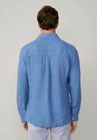 Camicia a maniche lunghe di lino azzurro chiaro con colletto con bottoni, due spalle posteriori e polsini con bottoni. Tessuto liscio, orlo leggermente curvo.