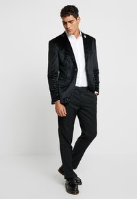 Blazer de terciopelo negro con un elegante brillo, cierre de botón único y una camisa blanca debajo. Combinado con pantalones oscuros y mocasines negros.