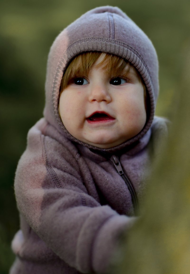 Hanorac cu fermoar din polar mov, cu glugă și manșete rib, purtat de un copil cu păr șaten deschis, zâmbind și ținându-se de un copac.