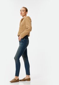 Chaqueta de textura marrón con dos bolsillos, llevada sobre jeans ajustados azules con detalles desgastados y mocasines marrones con un diseño texturizado.