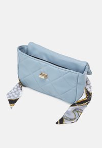 Ljusblått quiltat handväsk med ett guldspänne och ett mönstrat silkscarf knutet på ena sidan, visad mot en vit bakgrund.