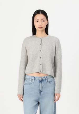 Grå kort cardigan laget av mykt materiale, med rund halslinning og seks frontknapper, båret over lyseblå høytlivsjeans.