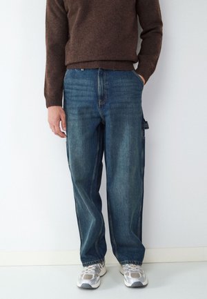 Man wearing a brown sweater, loose blue jeans with side pockets, and white athletic shoes standing against a plain white wall.
