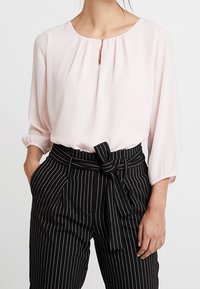 Ljusrosa blus med rund halsringning och nyckelhålsdetalj, i kombination med svartvita randiga byxor som har knytmidja och två fickor.