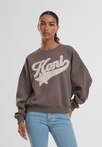 Felpa grigia con un logo "Kani" bianco invecchiato e design a stella. Tessuto morbido, vestibilità ampia, polsini e orlo a coste. Abbinata a jeans azzurri chiari.