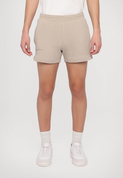 Beige Baumwollmix-Shorts mit elastischem Bund, ohne Taschen und einem dezenten Logo auf der linken Seite. Getragen mit weißen Sneakers und Socken.