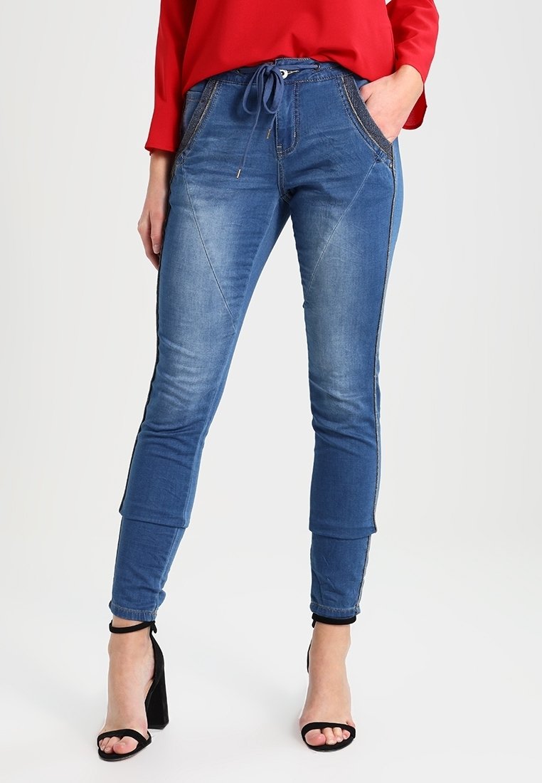 Pantalones jogger de mezclilla azul con cintura ajustable, corte ajustado y costuras laterales en contraste. Combinados con sandalias negras de tacón alto.