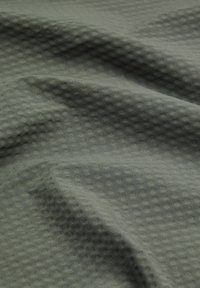 Texturerat grönt tyg med ett våffel-liknande mönster, mjukt och draperat, som visar en subtil glans och veck som indikerar flexibilitet.