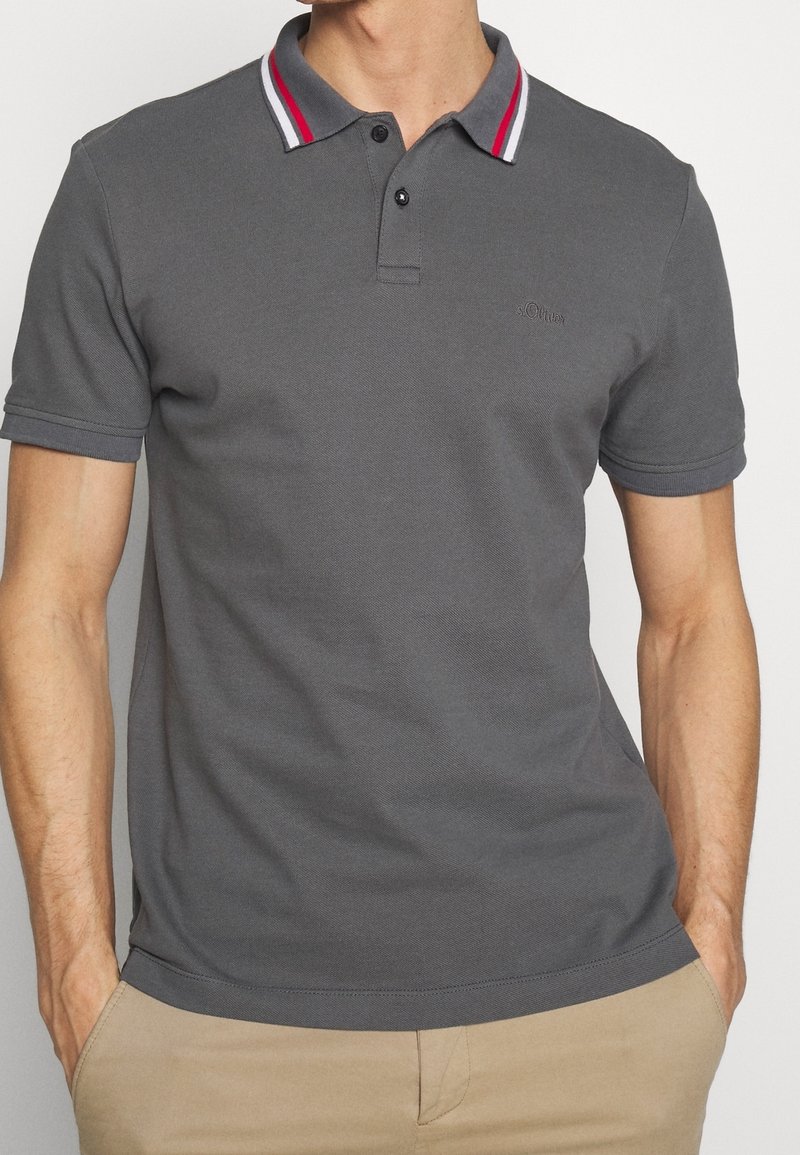 Homme portant un polo gris foncé avec un col rayé rouge, blanc et gris et un pantalon beige, les mains dans les poches, debout devant un fond uni.