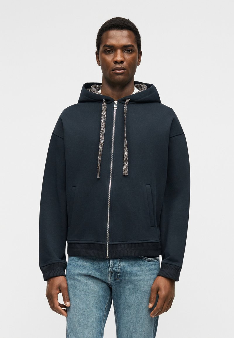 Homme portant un sweat à capuche zippé bleu marine avec des cordons à motifs et un jean bleu clair, debout devant un fond clair uni.