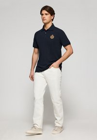 Polo marino con cuello y tapeta de tres botones, que presenta un logo bordado. Se lleva con pantalones blancos y zapatillas beige.