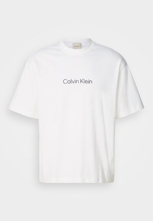 Biała bawełniana koszulka z krótkim rękawem, z szarym logo "Calvin Klein" umieszczonym na środku klatki piersiowej. Klasyczny okrągły dekolt, luźny fason.