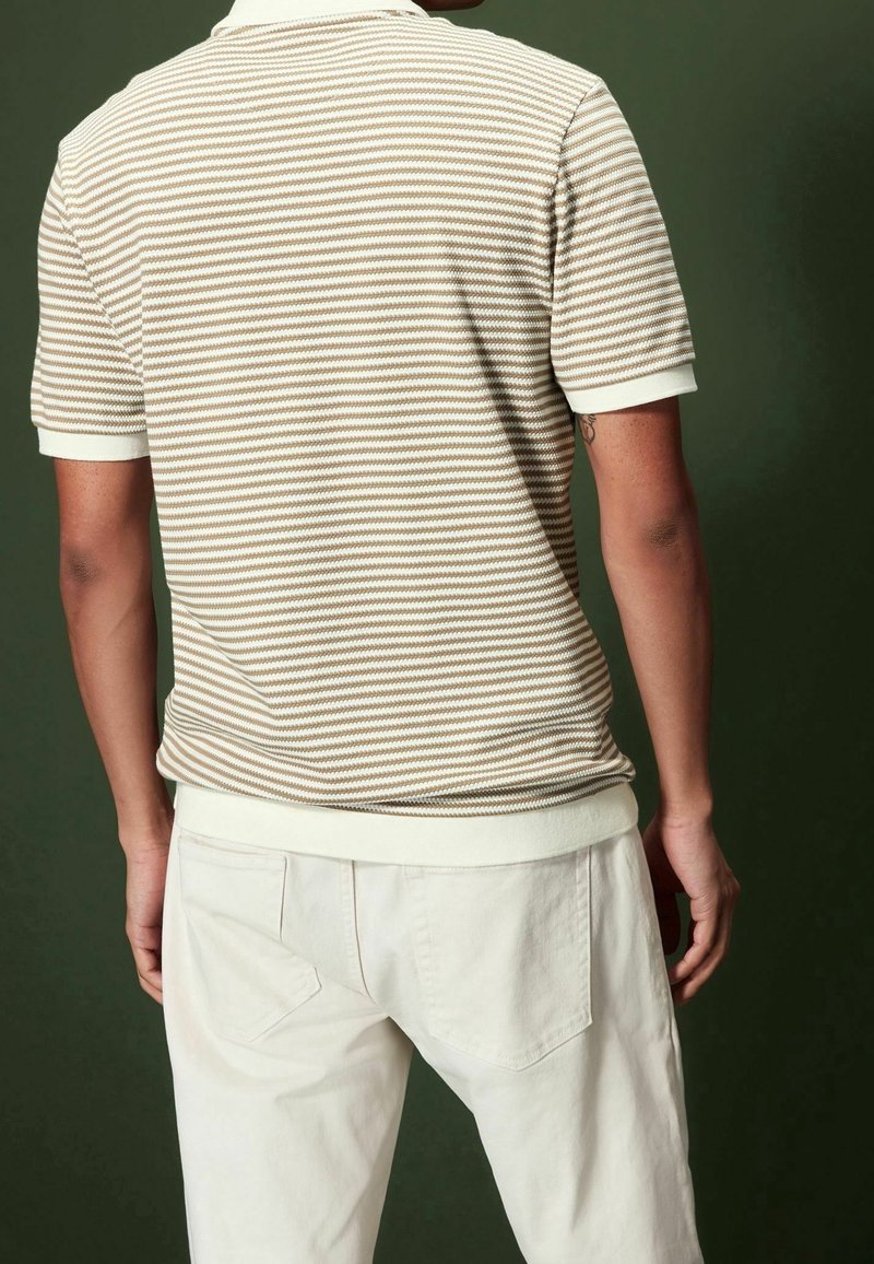 Beige und braun gestreiftes Polo-Shirt mit weißem Kragen, kombiniert mit cremefarbenen Hosen mit Gesäßtaschen. Strukturiertes Strickmaterial sichtbar.