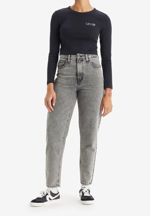 Persoon draagt een zwarte Levi’s top met lange mouwen, een hooggetailleerde vervaagde grijze jeans, witte sokken en zwart-witte sneakers, staand tegen een witte achtergrond.